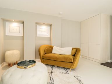 A living room with a mustard yellow sofa a white fur stool a round ottoman and built in wall shelves at The Hayloft in Sherston