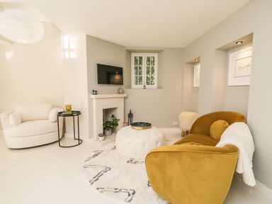 A living room with a mustard yellow sofa a white armchair a round ottoman and a wall-mounted TV at The Hayloft in Sherston