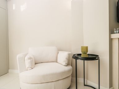 A white cushioned armchair next to a black round side table with a green glass on top in a corner at The Hayloft in Sherston