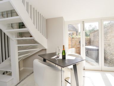 A dining area with a table set with a bottle and glasses next to a staircase and glass doors leading outside at The Hayloft in Sherston