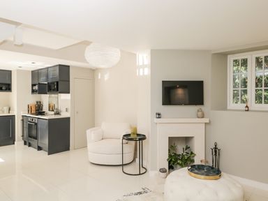 A kitchen area with black cabinets next to a living space with a white chair a side table a fireplace with plants and a mounted TV at The Hayloft in Sherston
