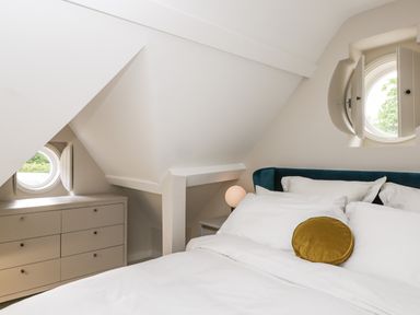 A bedroom with a bed white bedding a yellow round pillow a chest of drawers and round windows at The Hayloft in Sherston