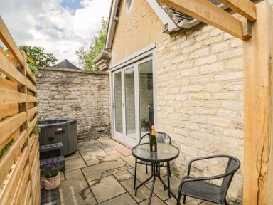An outdoor patio area with a glass table and two chairs a bottle and glass on the table a hot tub a potted plant and stone walls at The Hayloft in Sherston