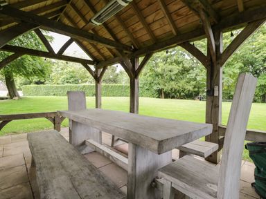 An outdoor wooden gazebo with a rectangular wooden table and benches surrounded by a grassy lawn at The Hayloft in Sherston