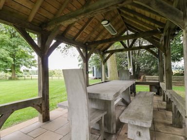 An outdoor wooden shelter with a table chairs and benches on stone tiles surrounded by grass and trees at The Hayloft in Sherston