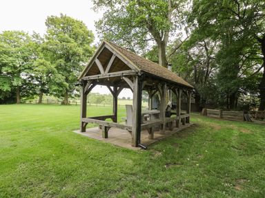 A wooden outdoor shelter with benches and a table on grass surrounded by trees at The Hayloft in Sherston