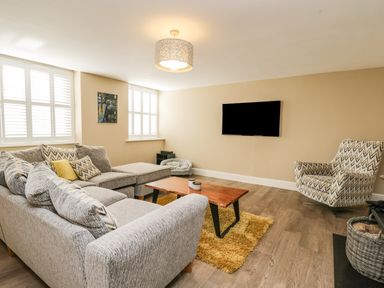 A living room with a grey sectional sofa a wooden coffee table a patterned armchair and a wall-mounted TV at The Coach House in Westonbirt near Tetbury
