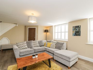 A living room with a grey sectional sofa, wooden coffee table, floor lamp, and windows with shutters at The Coach House Westonbirt near Tetbury