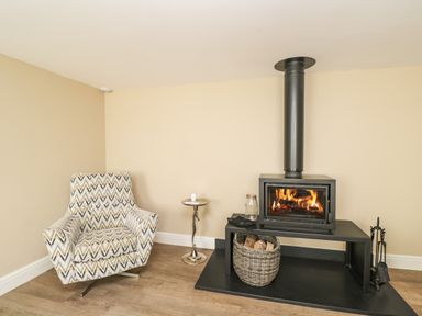 A corner with a patterned armchair next to a small round metal side table and a wood-burning stove with a basket of logs on a black hearth at The Coach House in Westonbirt near Tetbury