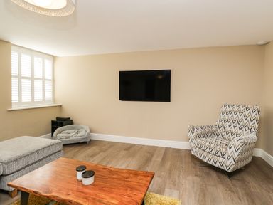 A living room with a wall mounted television a patterned armchair a grey ottoman a wooden coffee table and a dog bed at The Coach House in Westonbirt near Tetbury