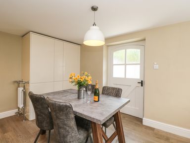 A dining room with a table and four chairs a vase with flowers a bottle and glass a cabinet and a door at The Coach House Westonbirt near Tetbury