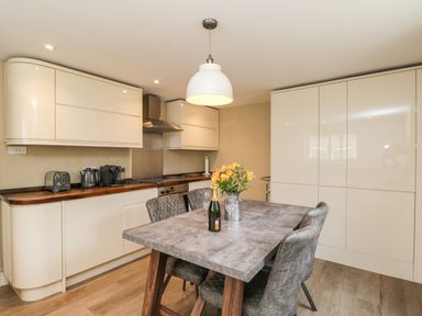 A kitchen with beige cabinets a wooden countertop a dining table with chairs flowers and a bottle at The Coach House in Westonbirt near Tetbury