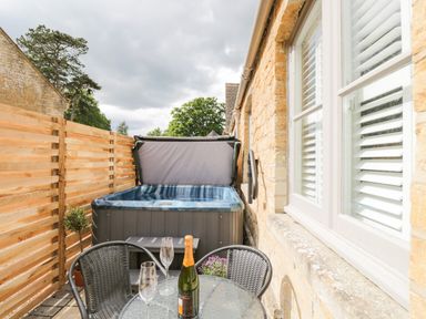 A small patio with a hot tub a round glass table with two chairs and a bottle with two glasses at The Coach House Westonbirt near Tetbury