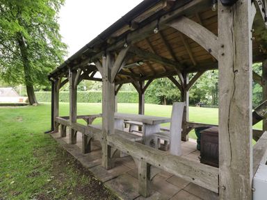 A wooden outdoor pavilion with table and chairs on a tiled floor in a grassy backyard at The Coach House Westonbirt near Tetbury