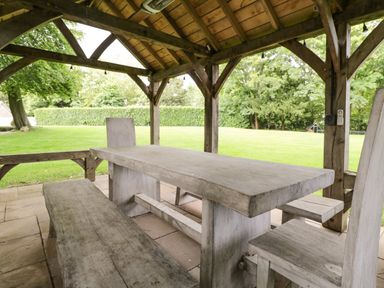 A wooden outdoor dining table with benches and chairs under a wooden gazebo in a garden at The Coach House in Westonbirt near Tetbury