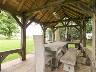 An outdoor covered seating area with a wooden table bench and high chairs on a stone floor at The Coach House in Westonbirt near Tetbury