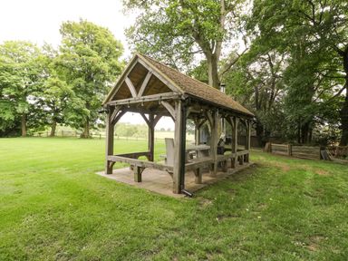 An outdoor wooden pavilion with benches and a table on a grassy area surrounded by trees at The Coach House in Westonbirt near Tetbury
