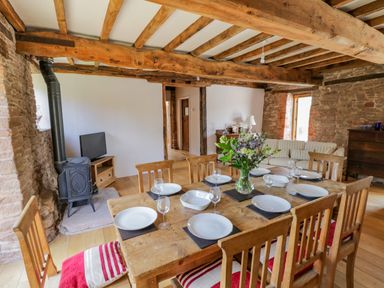 A dining area with a wooden table set with plates and glasses and a living area with a wood stove and a sofa at Munderfield Mill in Munderfield near Bishops Frome