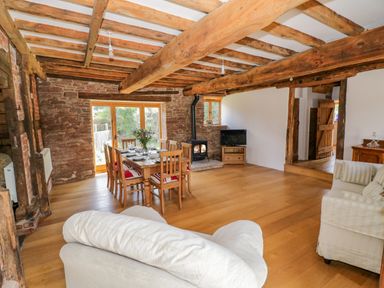 A dining area with a wooden table set for six and chairs a wood-burning stove a television and sofas with exposed wooden beams at Munderfield Mill in Munderfield near Bishops Frome