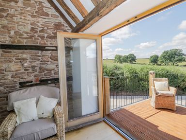An indoor seating area with a wicker armchair and cushions next to an open glass door leading to a wooden deck with another wicker chair overlooking fields at Munderfield Mill in Munderfield near Bishops Frome