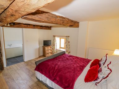 A bedroom with a bed covered by a red blanket and pillows wooden beams a window with floral curtains a wooden chest of drawers with a TV and an adjoining bathroom at Munderfield Mill in Munderfield near Bishops Frome