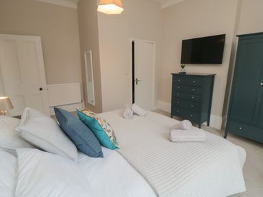 A bedroom with a bed with pillows and towels a chest of drawers with a plant a wall-mounted TV and a wardrobe at Spinnaker House in Scarborough