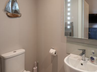 A bathroom with a toilet, wall-mounted paper holder, sink with faucet, hand wash bottle, mirror and metal sailboat wall decor at Spinnaker House in Scarborough