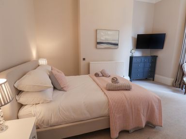 A bedroom with a bed covered with white and pink bedding towels on the bed a lamp on the bedside table a chest of drawers with a vase of flowers and a wall-mounted TV at Spinnaker House Scarborough