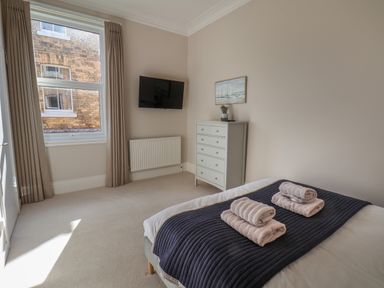 A bedroom with a bed, folded towels, a dresser with a plant, a wall-mounted TV, and a window with curtains at Spinnaker House in Scarborough
