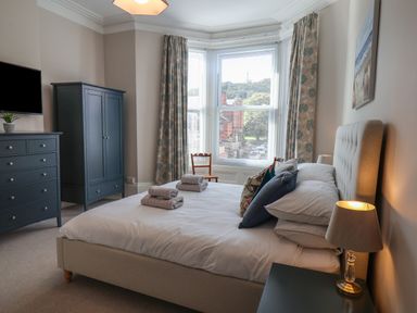 A bedroom with a bed, bedside table with lamp, chest of drawers, wardrobe, two chairs by a bay window with curtains at Spinnaker House in Scarborough