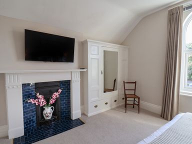 A bedroom corner with a mounted TV above a fireplace with flowers a white wardrobe a wooden chair and a window with curtains at Spinnaker House in Scarborough