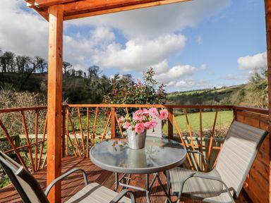 A covered wooden balcony with a glass table and two chairs overlooking fields and trees at Bearscombe View in Modbury