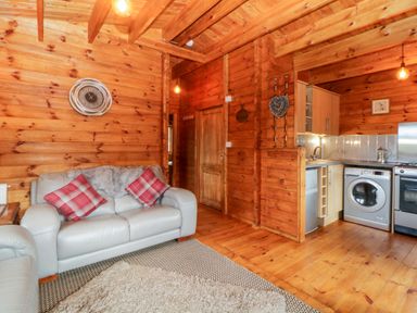 A wooden living room with a light gray sofa with red plaid cushions a kitchen area with a washing machine fridge and oven and a wall clock at Bearscombe View in Modbury