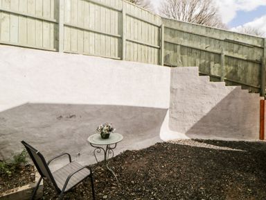 An outdoor area with a metal chair and round table with flowers against a white retaining wall and wooden fence at Bearscombe View in Modbury