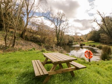 A wooden picnic table on grass near a pond with trees and a lifebuoy at Bearscombe View in Modbury