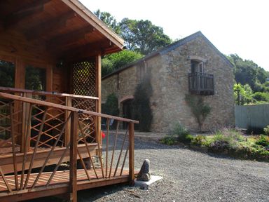 A wooden porch with railing next to a stone building with a small balcony surrounded by plants and trees at Bearscombe View in Modbury