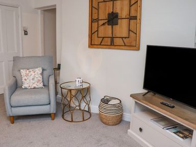 A sitting area with a gray armchair with a floral cushion next to a round glass table a basket with blankets and a television on a wooden stand at Monteva Whitby