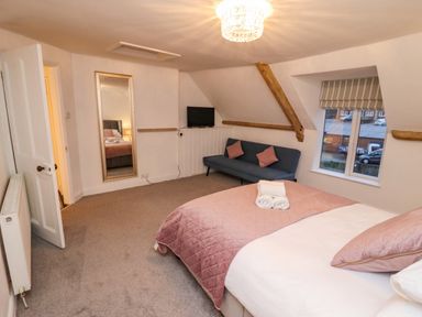 A bedroom with a double bed covered with a pink quilt and pillows a blue sofa with pink cushions a wall-mounted television a tall mirror and a window at Monteva Whitby