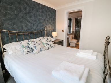 A bedroom with a metal bed frame white bedding patterned pillows bedside table and lamp and an en suite bathroom at Mayflower in Broadwoodwidger near Ashwater