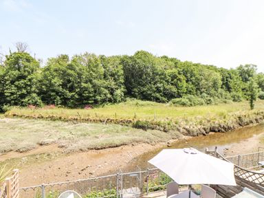 A small river with a grassy bank and trees beyond a fenced patio area with a table and umbrella at Penpoll in Little Petherick Creek near Padstow
