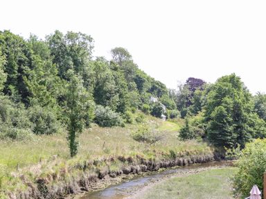 A river running alongside a grassy bank with trees and a white house in the background at Penpoll Little Petherick Creek near Padstow