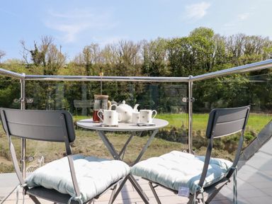 A balcony with a round table set for tea and two chairs with cushions overlooking trees and grass at Penpoll in Little Petherick Creek near Padstow