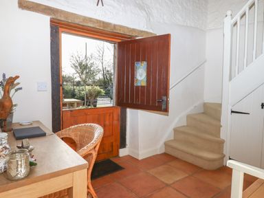 A hallway with an open wooden door a wicker chair a wooden table and carpeted stairs at The Old Barn in Colyton
