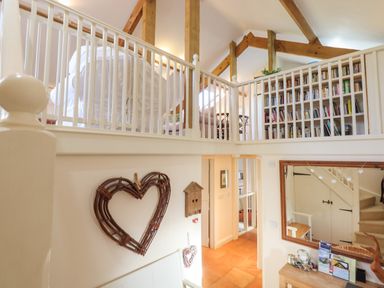 An interior view of a two-level room with wooden beams a white railing a large bookshelf and a heart-shaped wall decoration at The Old Barn in Colyton