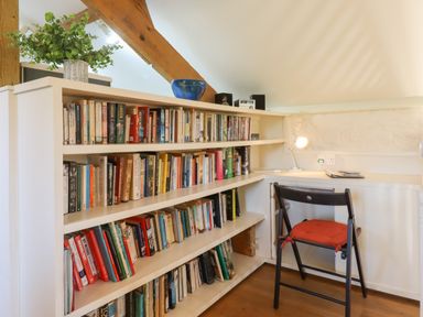 A study corner with bookshelves filled with books a wooden chair with a red cushion and a small desk with a lamp at The Old Barn in Colyton