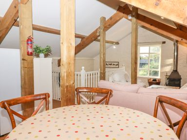 A dining area with a round table and wooden chairs next to an open living space with sofas wooden beams and a wood stove at The Old Barn in Colyton
