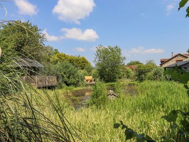A garden with a pond surrounded by tall grass, trees, a wooden gazebo on the left, a bench in the background, and stone buildings at The Old Barn in Colyton
