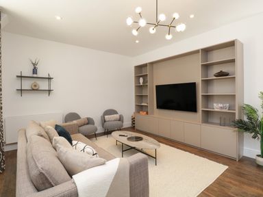 A living room with sofa, armchairs, coffee table, TV on a shelving unit, wall shelves, and plant at Alice House Stone Cross Mansion in Ulverston