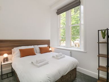 A bedroom with a bed with white linens and orange pillows a wooden headboard a window and a shelving unit with a plant at Alice House Stone Cross Mansion in Ulverston