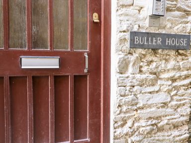 A brown wooden door with a mail slot and a lock next to a stone wall with a sign reading Buller House at Smugglers Loft in Looe